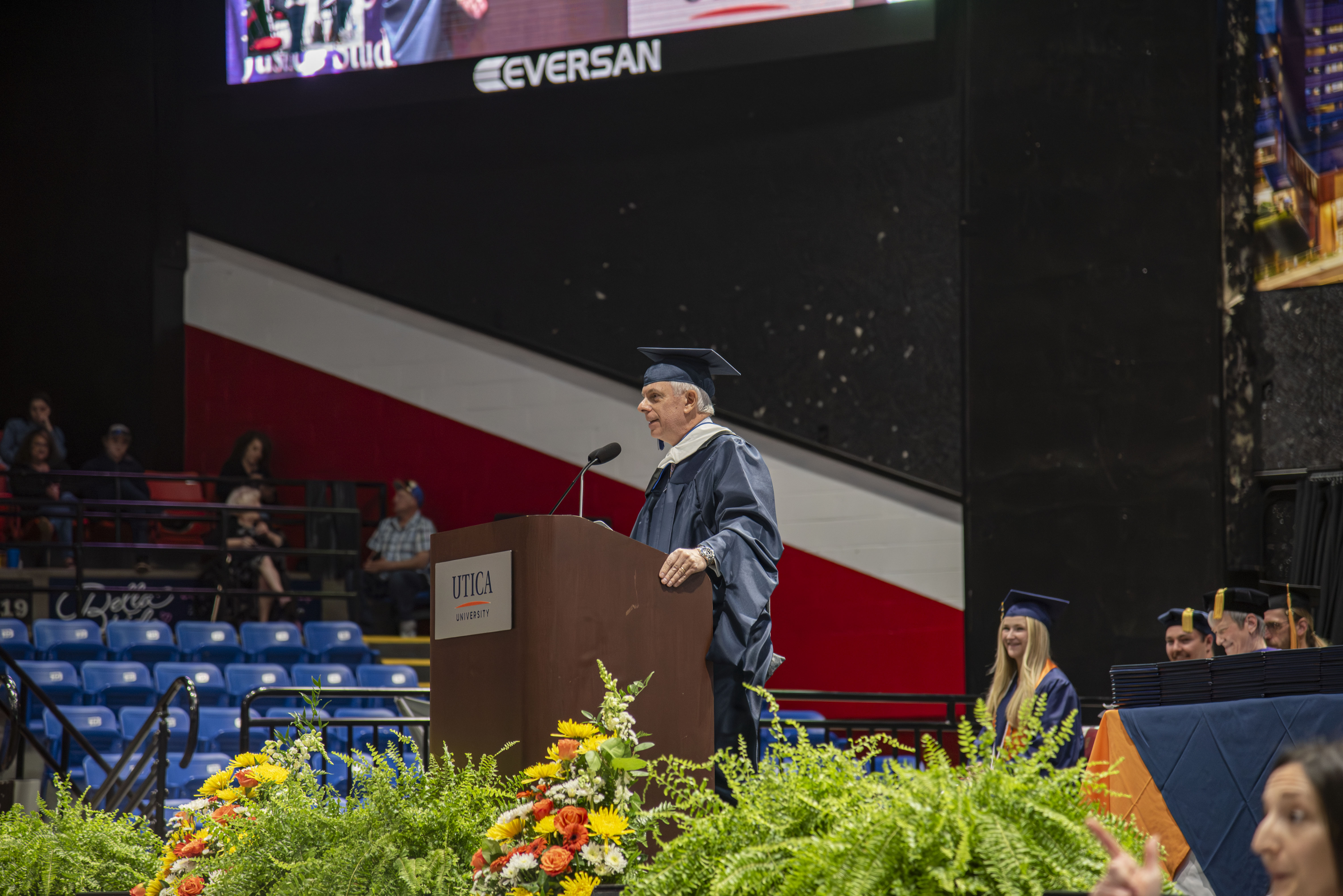 Anthony Picente, in blue gown and cap, stands at a lectern and addresses the graduates of 2026.