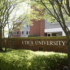 The words Utica University against a brick wall in front of a residence hall.