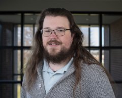 Tristan Morgan in glasses and gray sweater stands in front of windows.