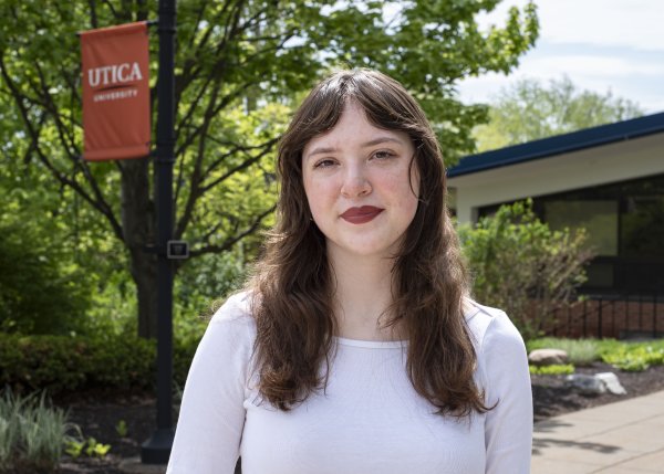 Bella Stephens, in white top with brown hair down past shoulders, stands in front of foliage on campus.